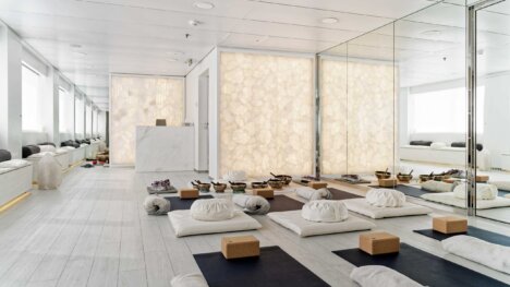 dampa sustainable ceiling applied in a white yoga studio featuring a crystal wall and a mirror wall one one side with a wall with windows and seating on the other side. in the middle of the room are many yoga mats, yoga blocks, towels and cushions.